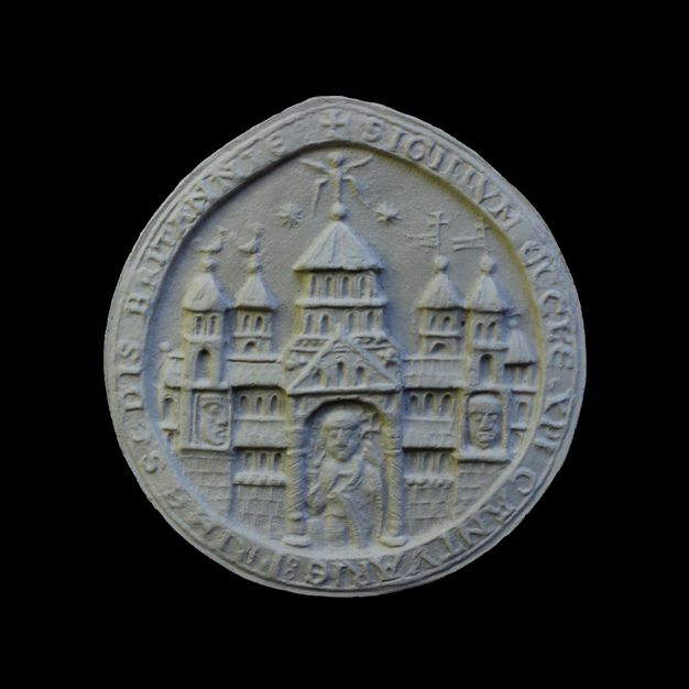 Seal. Cast of a round seal shows a church with five towers over a porch with a triangular pediment. A figure stands beneath the pediment. Two faces, one to each side, are in niches under towers at either end of the building. Latin text lines the perimeter.
