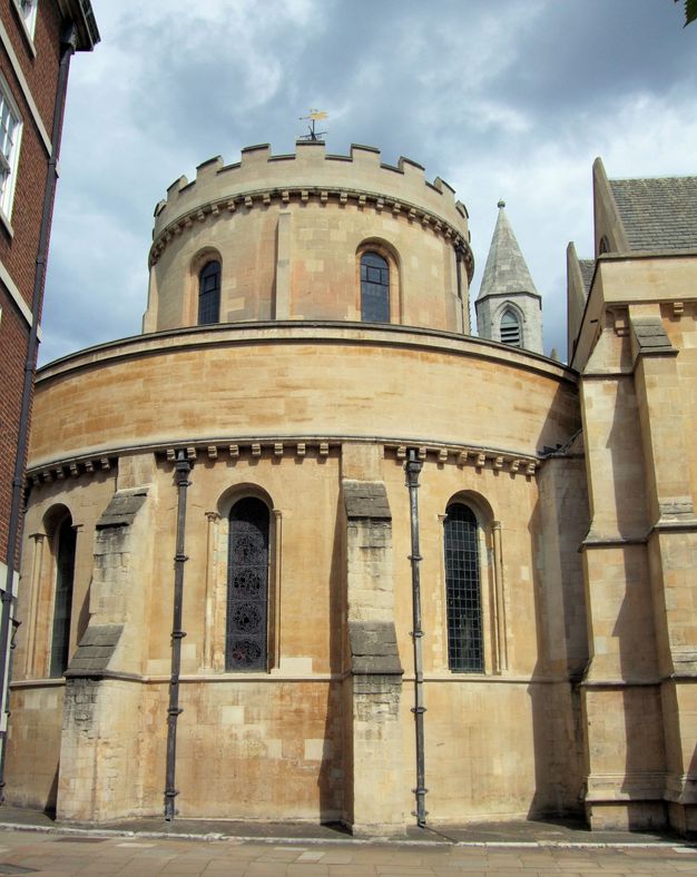 Architecture. A round church made of cream coloured stone is tucked between other buildings. Engaged buttresses alternate with narrow, arched windows. The narrower, also circular, second storey has crenelations around the top edge.