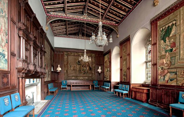 Hall-like room is decorated with carved brown wood moulding and panelling, a timbered and painted ceiling, stained glass windows, two chandeliers, and tapestries.