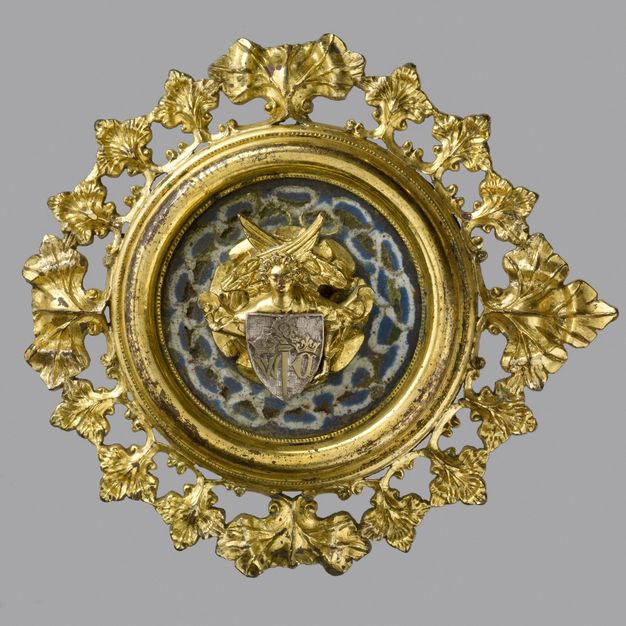Plaque. A golden, medallion-like roundel has a gold angel with crossed wings and a silver-coloured shield at its centre, against a background enamelled in blue with a white figure-of-eight pattern. The perimeter is lined with flaring gold leaves.