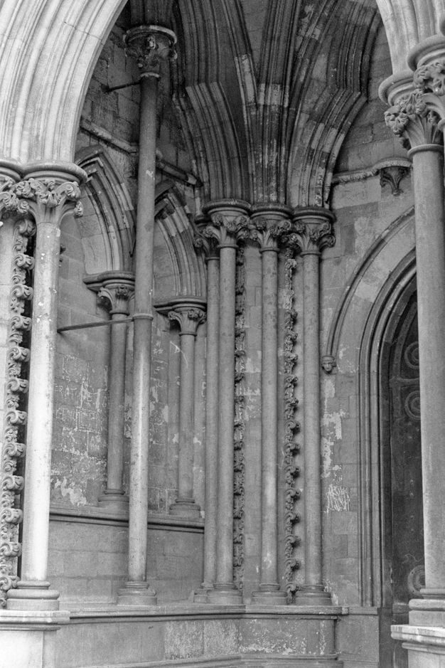 Architecture. Black and white photograph of slender stone columns clustered in the corner of a church porch hold up tri-lobed arches so layered they resemble phyllo dough. A detached column stands far out in front of the blind arches to our left.