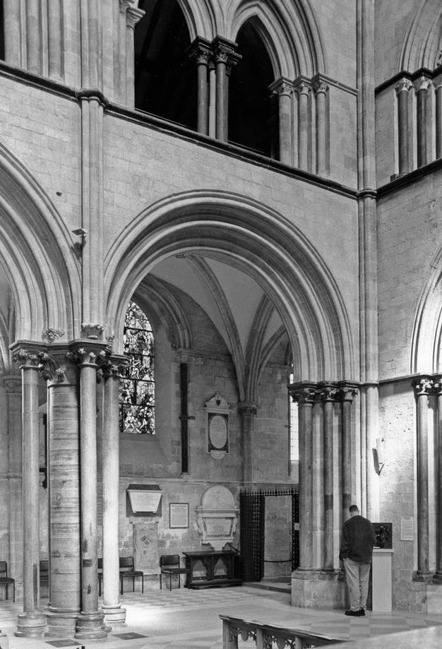 Architecture. Black and white photograph of a wide, rounded arch supported on clustered columns and opening onto a shallow chapel. The double arched opening of the gallery level above is in deep shadow.