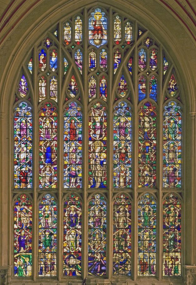 Stained glass window. Seven tall columns of glass under a pointed arch are subdivided by overlapping arches across its width. Scenes with people fill every sliver of space with vibrant, jewel-toned colour.