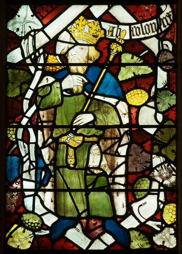 Stained glass window. Detail view of a king wearing a crown and green robe under a fur-lined cape. He holds a sceptre in one hand and strokes his long beard with his other. A scroll near his head has Gothic writing that is difficult to make out.