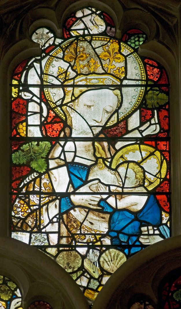 Stained glass window. Detail view of a crowned woman, the Virgin Mary, nursing a baby, Jesus. Her breast is near her left shoulder, to our right. Both have halos, and the spaces around them are filled with a red leafy pattern.