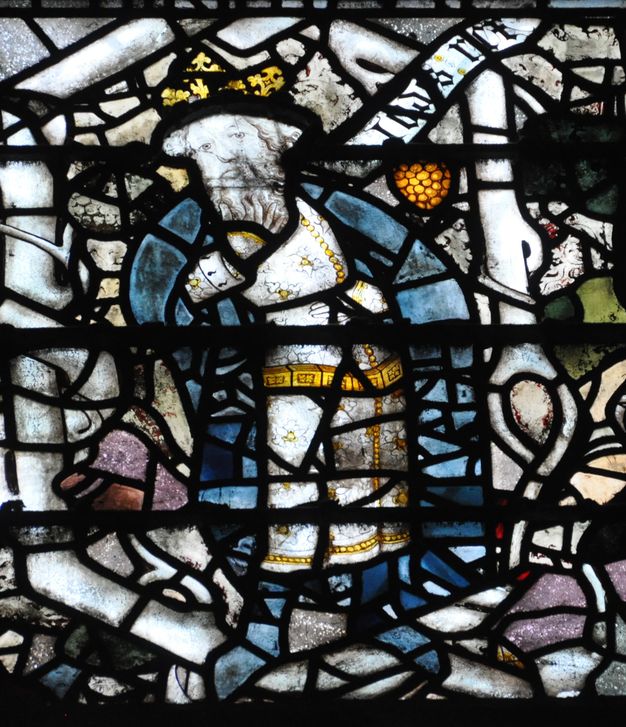 Stained glass window. Detail view of a king wearing a crown and white tunic under a blue cape. He rests one hand against his belly and strokes his long beard with the other. A scroll near his head has Gothic writing that is difficult to make out.