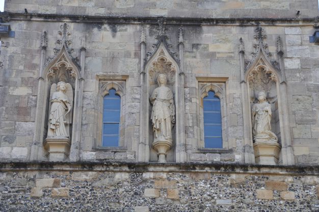 Architecture. Three niches spaced across a stone wall show a woman, the Virgin Mary, in the centre and a winged angel, Gabriel, in the niche to our left. A man kneels in the rightmost space. Each niche is lined with tracery under triangular peaks above, which are lined with flame-like decorations.