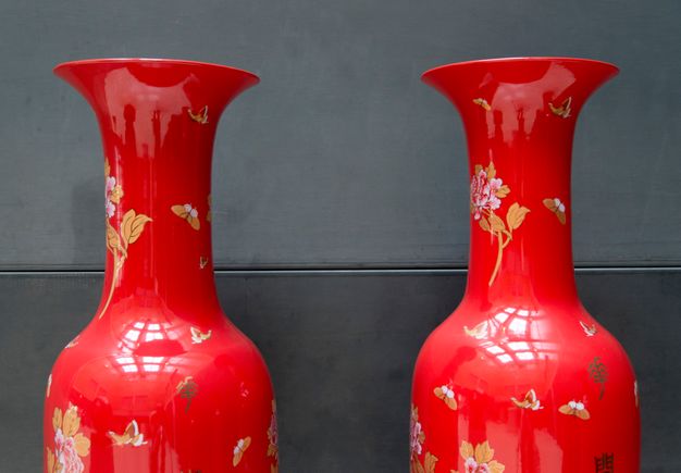 Installation photograph. The vibrant red shoulders, long necks, and flaring mouths of two ceramic vessels reflect the light in the room and the photographer taking this picture. Butterflies and flowers in shades of golden tan, white, and pale pink are scattered across the vessels’ surfaces.