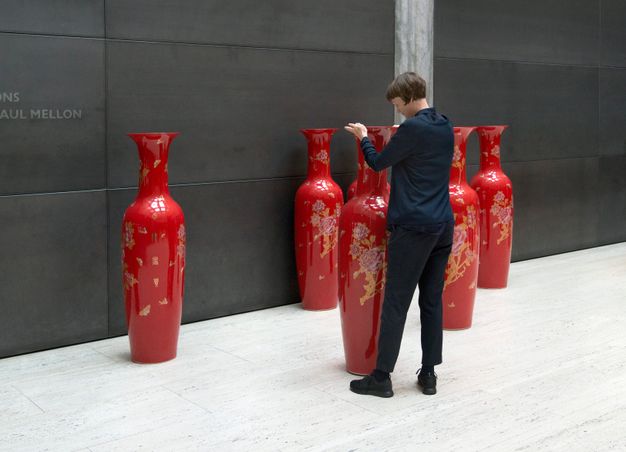 Installation photograph. A woman adjusts the placement of a slender vibrant red vase that comes up to her shoulder height. Five more vases are along or near a black wall opposite us.