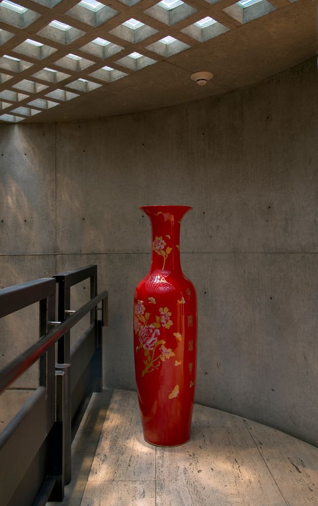 Installation photograph. A vibrant red vessel stands in a curving corner near a railing. The vessel has a flaring mouth, a slender neck, and a tall, narrow body. Leaves and butterflies in gold, pink, and white are painted on the vessels. Dappled light from the grid-like skylight falls across the vessel and stone floor.