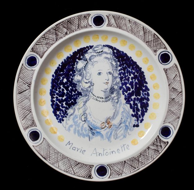 Plate. A woman looks off to our right. Her shoulders are covered with dashes and squiggly strokes, suggesting ruffles. The woman and the dotted background are painted mostly in shades of blue with a couple of touches of brown. A ring of yellow dots runs just inside the rim of the plate, and her name is painted at the bottom centre, \“Marie Antoinette\”.