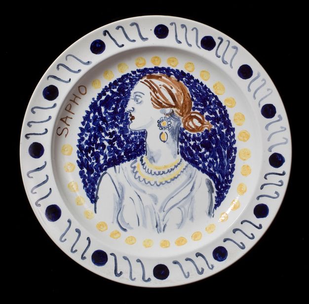 Plate. A sketchily painted woman turns her face so we see her in profile even though her shoulders are square to us. She and the background are painted mostly in shades of blue, and her brown hair is pulled back into a low bun. A ring of yellow dots runs just inside the rim of the plate, and her name is painted to the left, \“Sapho\”.