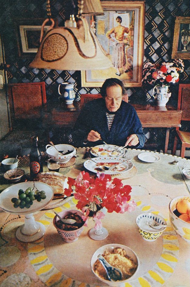Colour photograph. A balding man with dark hair sits at a round table, eating. Dishes and platters are spread across the table. Paintings hang on the far wall against a dark, geometric pattern of black, grey, and gold.