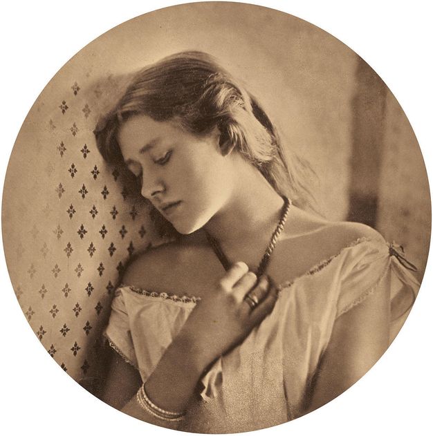 Sepia-toned photograph. Shown from the chest up, a pale-skinned woman leans her head against a wall and looks down in this circular portrait. She touches her necklace with one hand. Her hair falls down her back and a loose white garment sits low on her shoulders.