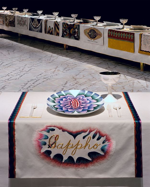Installation. A place setting at a long table has a plate painted with a pink, blue, purple and green flower over a section of tablecloth embroidered with gold text that reads \“Sappho\”. The S has a harp as the left side of the letter. Other place settings line another table angling away from us.