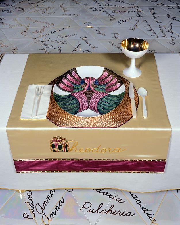 Installation. A place setting at a table has a plate painted to look like a mosaic in a gold, magenta, blue, and green. The plate and a gold-lined white goblet sit on a pale gold cloth embroidered in darker gold, reading \“Theodora\”. The capital T is overlaid on an architectural arch.