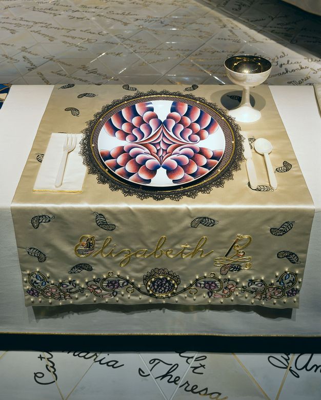 Installation. A place setting at a table has a plate painted with red and pink, lobe-like forms extending from a clustered centre to the edges of the white plate. The gold cloth beneath this setting is embroidered in darker gold thread, \“Elizabeth R\”.