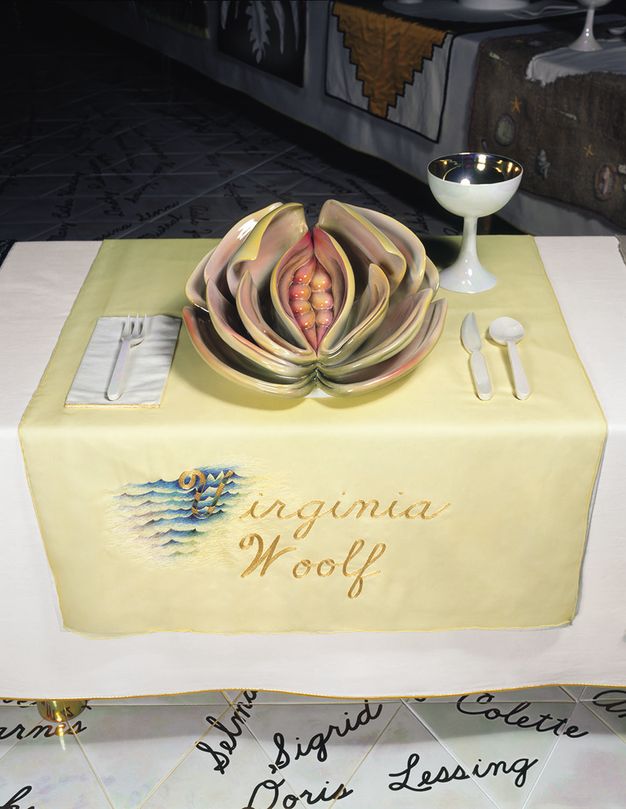 Installation. A place setting at a table has a highly sculptural plate with deep folds around a narrow opening in shades of pale pink, yellow and green. The yellow cloth beneath this setting is embroidered with gold letters, \“Virginia Woolf\”.