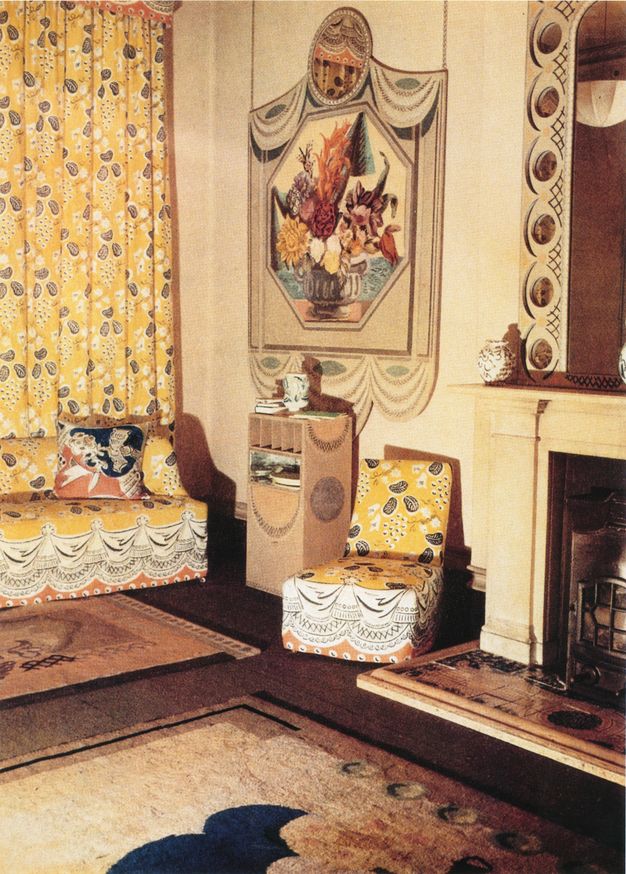 Colour photograph. A couch and an armless chair in the corner of a room are upholstered in a pattern of soft black and white shapes on a canary-yellow background. The same fabric is used for the floor-length curtains behind the couch. A floral still life is on the wall behind the chair, next to the neighbouring fireplace, and there are rugs on the floor.