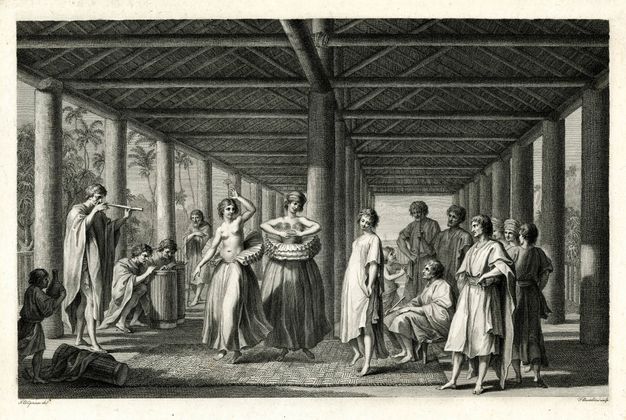 Print. Two women dance as two men play drums and a third man plays a pipe. Both women wear voluminous ruffs around their waists over long, full skirts and bare feet. One woman wears a turban and a gauzy material over her breasts. The other is bare-headed and bare-chested. They and several onlookers are under a timbered roof held up with columns. Palm trees are visible in the distance. 