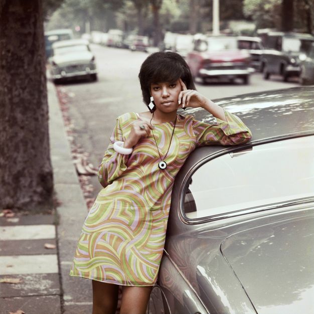 Colour photograph. A young black woman wears a long-sleeved sheath dress patterned in muted green, yellow, brown, and pale purple. On a car-lined street, she leans against a shiny grey car, one elbow resting on the hood with her hand against her face.