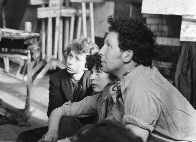 Black and white photograph. Two men and a woman look up and to our left in a studio. They are close to us so are shown from the waist up. The man and woman closest to us have dark hair, and the third has lighter curls.