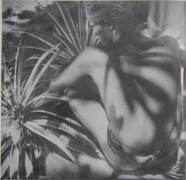 Black and white photograph. Shadows cut across the back of a dark-skinned man seated facing away from us with his arms around his knees. He wears a cloth wrapped around his waist and another around his head. Spiky tropical plants fill the space around him.