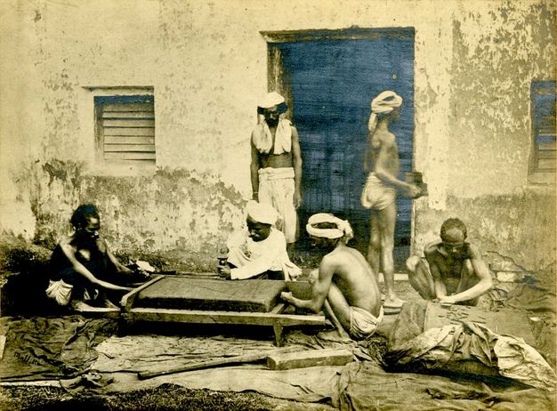 Black and white photograph. Three men work around a platform on the ground in front of a building façade with peeling paint. Three others crouch or stand around the platform. The men have dark skin and are thin and wiry. Four wear white cloths around their heads, and their clothing ranges from loincloths to a long-sleeved tunic. The door and two windows of the building are coloured indigo blue.