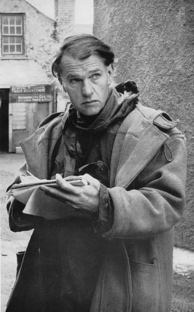 Black and white photograph. Shown from the hips up, a man with a long face and square chin looks up and off to our right, his brow knit. He wears a long, heavy coat and holds a sketchbook in one hand.