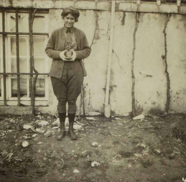 Black and white photograph. A woman smiles at us as she holds a camera in front of her torso. She wears a jacket, button-up shirt, and jodhpur-like trousers over ankle boots.