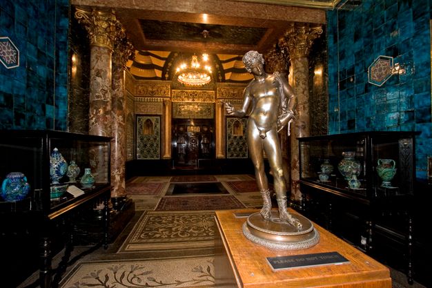 Colour photograph. A gleaming bronze sculpture of a man, about half size, stands on a table in a hall-like room with mosaic floors, tiled walls, and stone columns with gold capitals. Patterns abut patterns throughout the space.