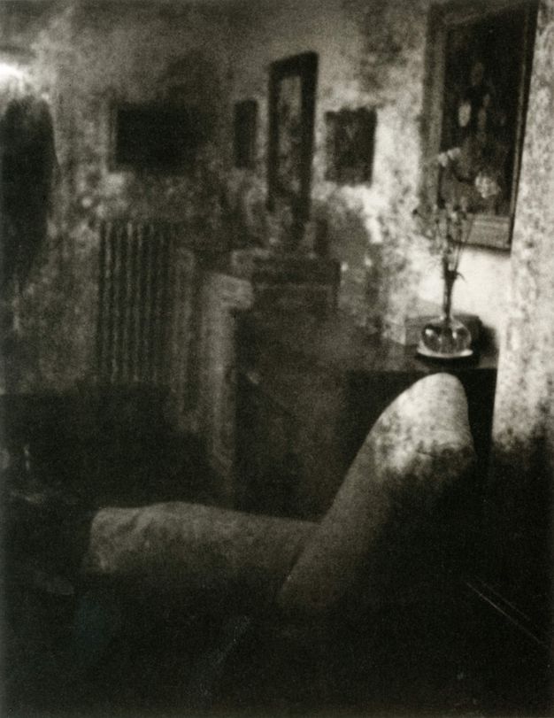 Black and white photograph. A blurry room is shown room with an upholstered chair, side table, paintings, and a radiator. The blurry quality of the photo creates a dreamlike haze.