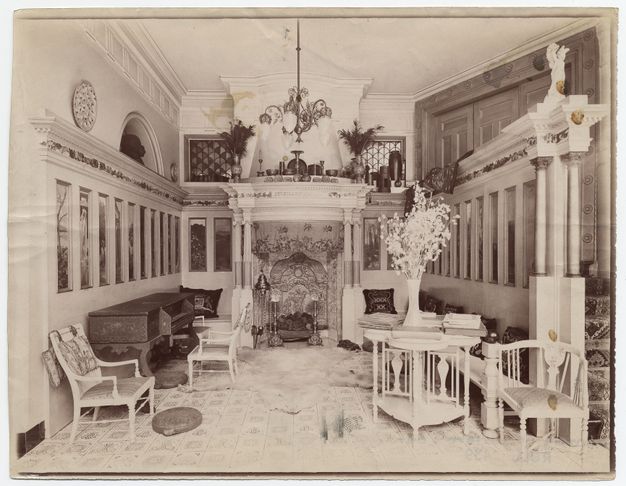 Black and white photograph. A hall-like room is lined with tall, narrow panels under an architectural entablature leading to an ornate mantlepiece over a richly decorated fireplace across from us. The floor is also patterned, and delicate white furniture is arranged along the edges of the space.