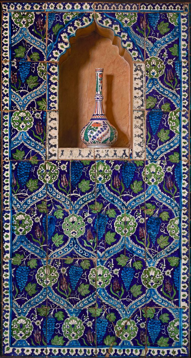 Architecture. Wall tile makes an overarching pattern of continuous S’es surrounding grapes, leaves, and flowers. A blue, white, green, and dark orange vase with a long, narrow neck and flaring body sits in an arched niche near the top.
