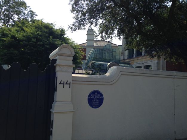 A round blue sign on a white garden in blue text. Trees and rooflines fill the space beyond the wall.