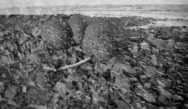 Black and white photograph. Rocks leading back to surf nearly fill this composition. Another image, perhaps of more rocks, is a ghostly echo superimposed on the landscape.