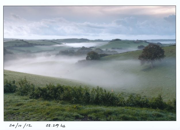 Thick, seemingly tangible fog pools in valleys that meander back through rolling hills in this landscape. Handwritten text under the photograph provides the date and time.