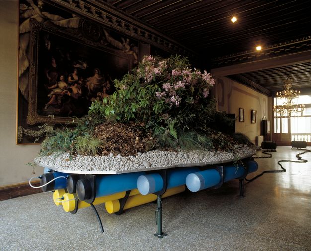 Installation. A bank of rhododendrons sits on a mound of dirt supported by a layer of white rock. Blue and yellow pipes run under the tray holding the plants. The piece is set up in a gallery with wood ceilings, a brass chandelier, and stone floors.
