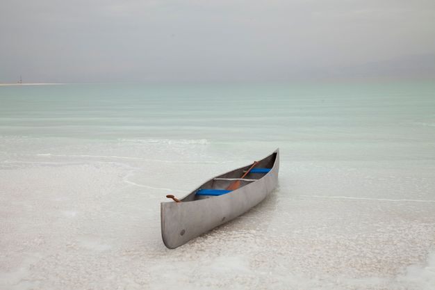 A silver-coloured canoe sits in the shallows of water that deepens from ivory white close to us to pale glacial green in the distance. The horizon is hazy lavender blue.