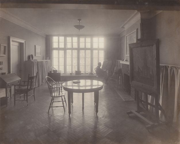 Black and white photograph. A round table and one chair is backlit by a wall of windows at the far end of this room. A desk, easel, and other furniture lines the walls. A sitting person is silhouetted near the windows.