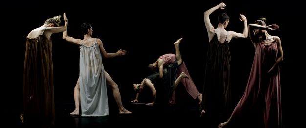 Six dancers wearing long, flowing gowns pose with arms and legs flexed or rigid against a black background.