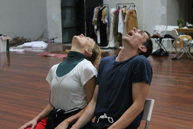 Colour photograph. A man and a woman sit side by side in a rehearsal space. Both throw their heads back so we look on the undersides of their chins. The man’s mouth is wide open.