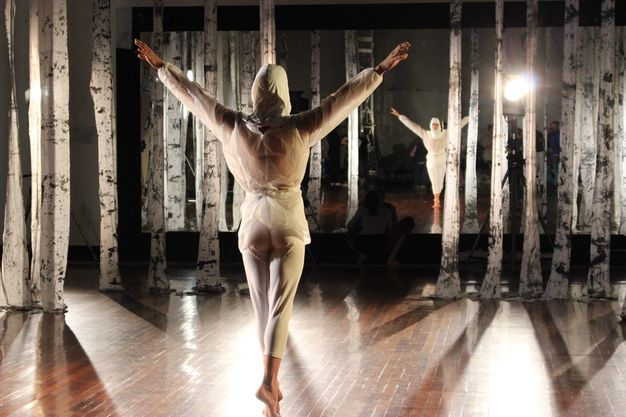 A dancer balances on his tip toes, both arms stretched out and up. He wears a white hood and clothing. He faces away from us and is reflected, along with the filming crew, in a mirror on the wall opposite us. Strips of white fabric hanging from the ceiling are streaked with dark grey.
