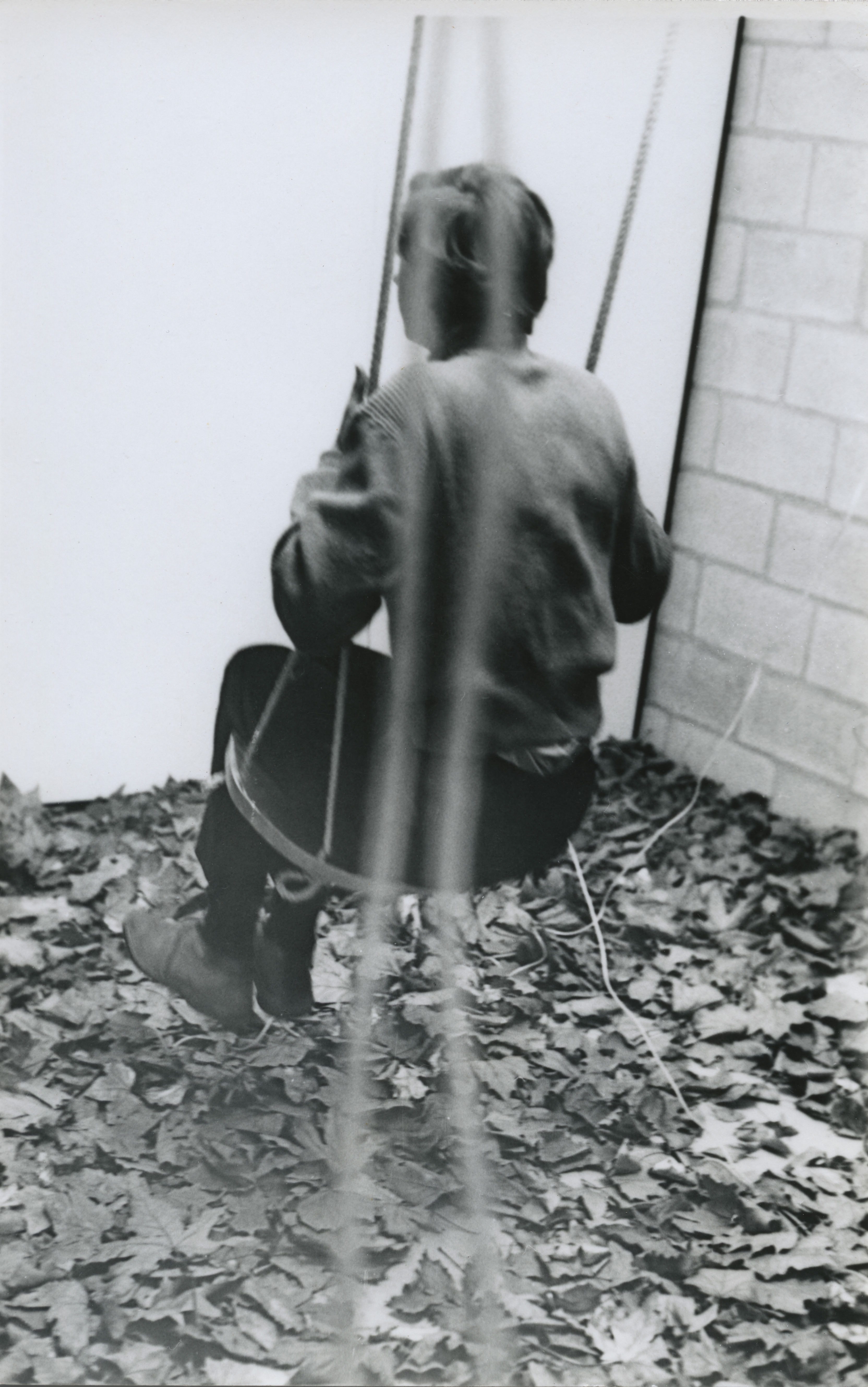 Black and white photograph. A person swings away from us near a corner. The ground is covered in leaves.