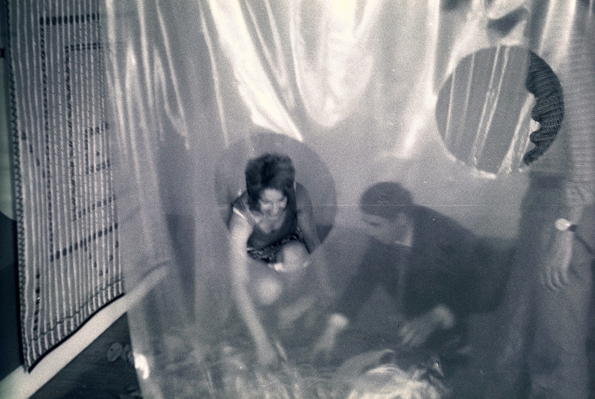 Black and white photograph. Holes are cut out of a sheet of translucent plastic hanging between us and visitors beyond. A man and a woman crouch and both reach for something on the floor.