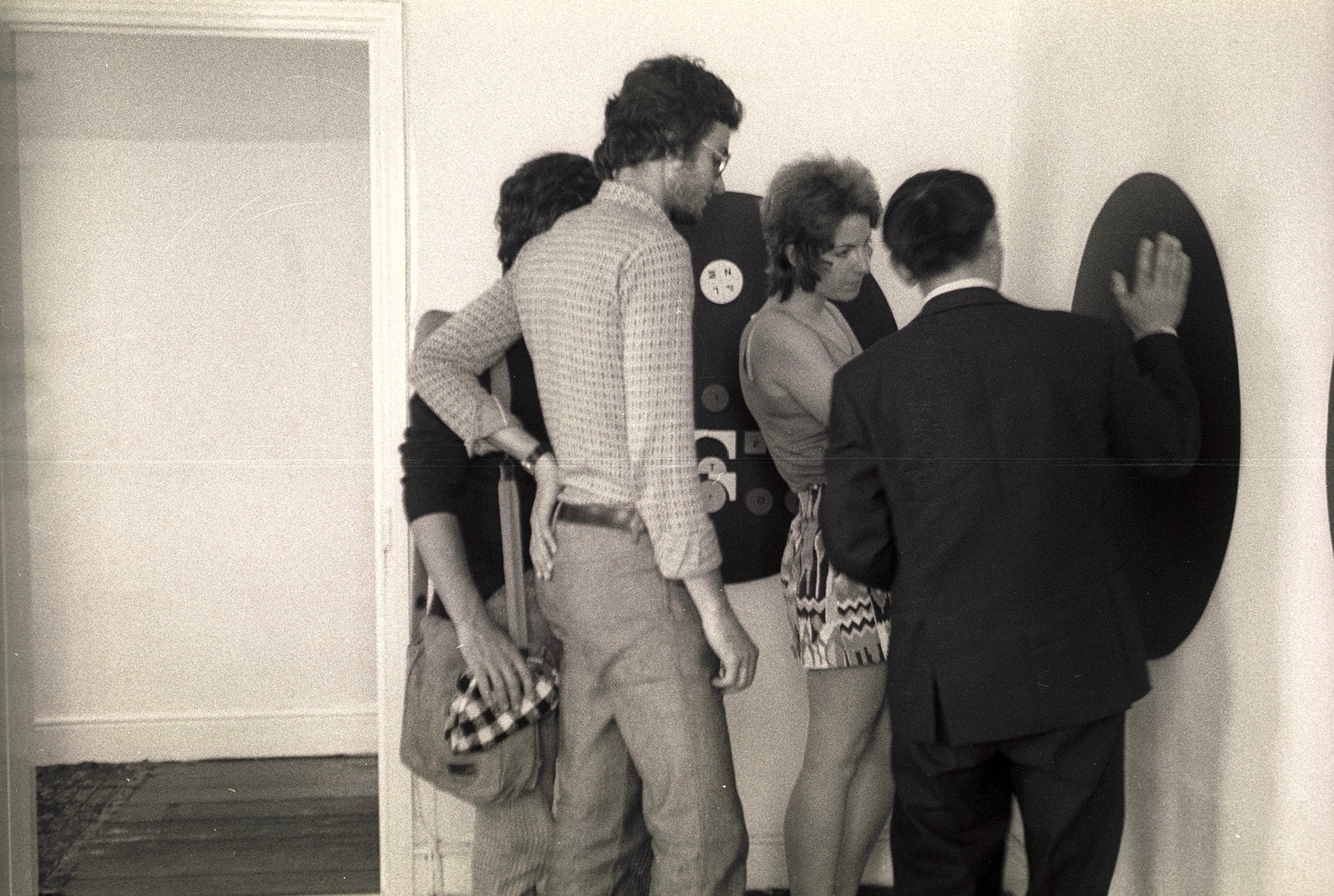 Black and white photograph. Four people stand at a black disc, about half the height of the people, on a gallery wall. A person facing away from us, wearing a suit, gestures towards the disc.