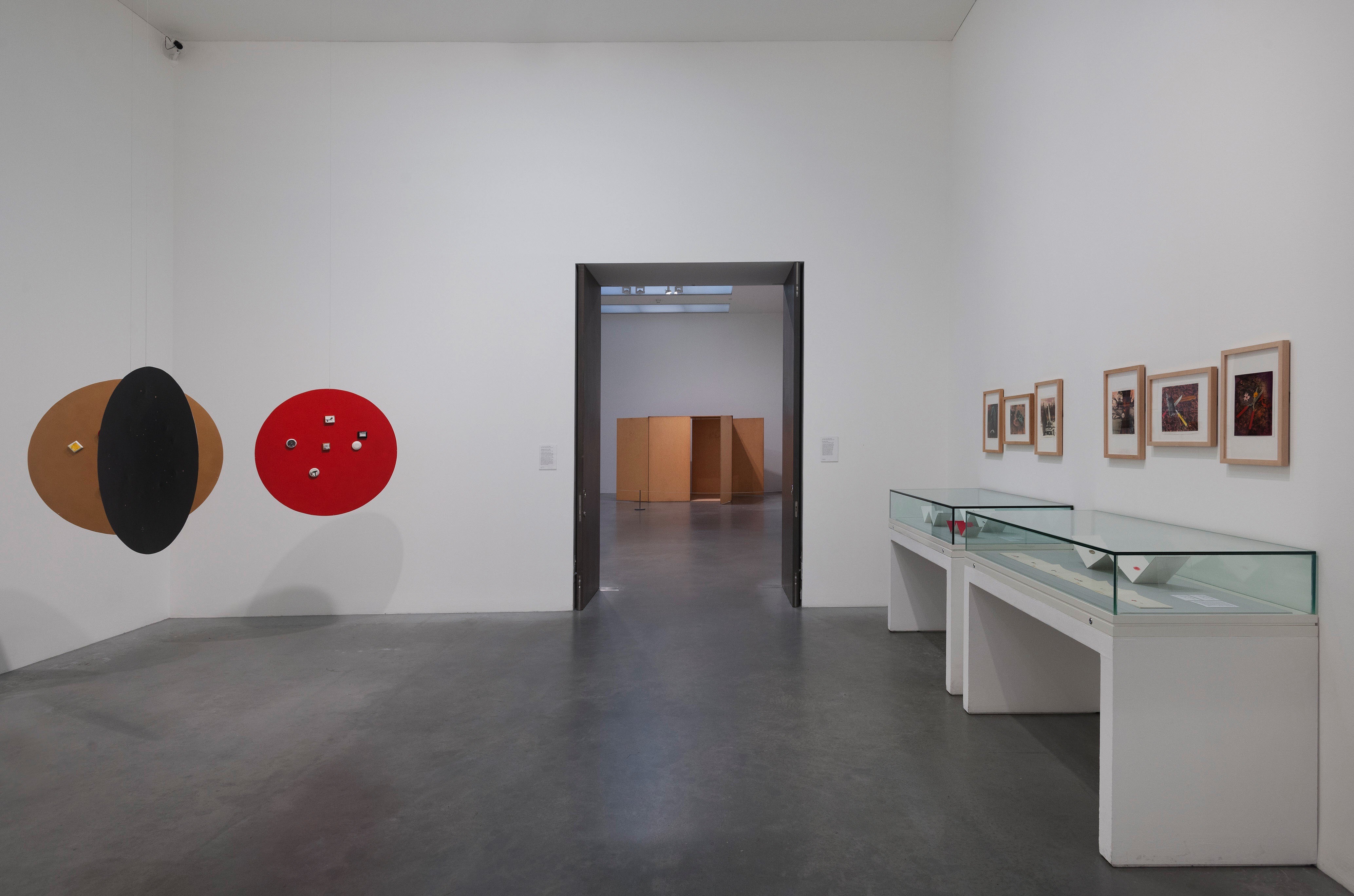 Installation. To our right, six framed works of art hang over two long cases with accordion-folded strips of paper in this gallery. Three discs, one red, one black, and one fawn brown, are suspended from the ceiling to our left.