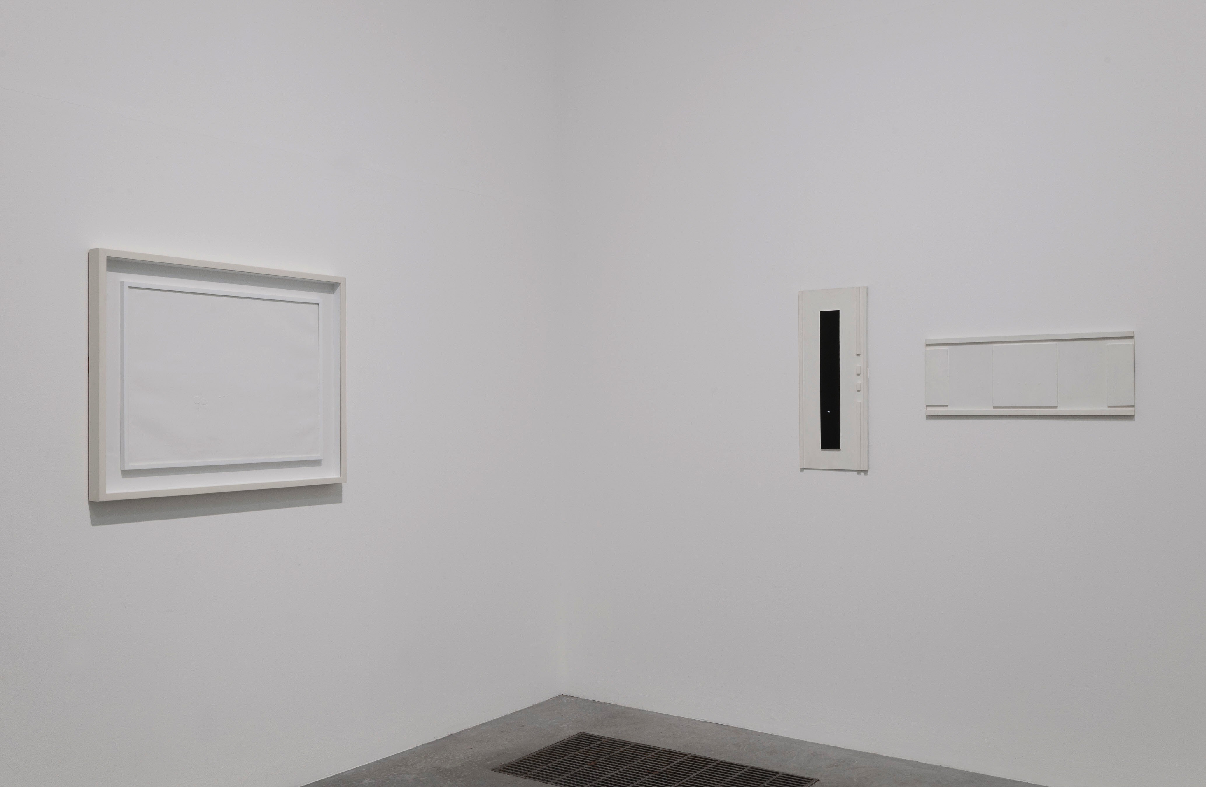 Installation. Three works of art are hung in the corner of a gallery. A rectangular piece to our left and a long, narrow piece to our right are totally white. The third has a black stripe down a white support or background.