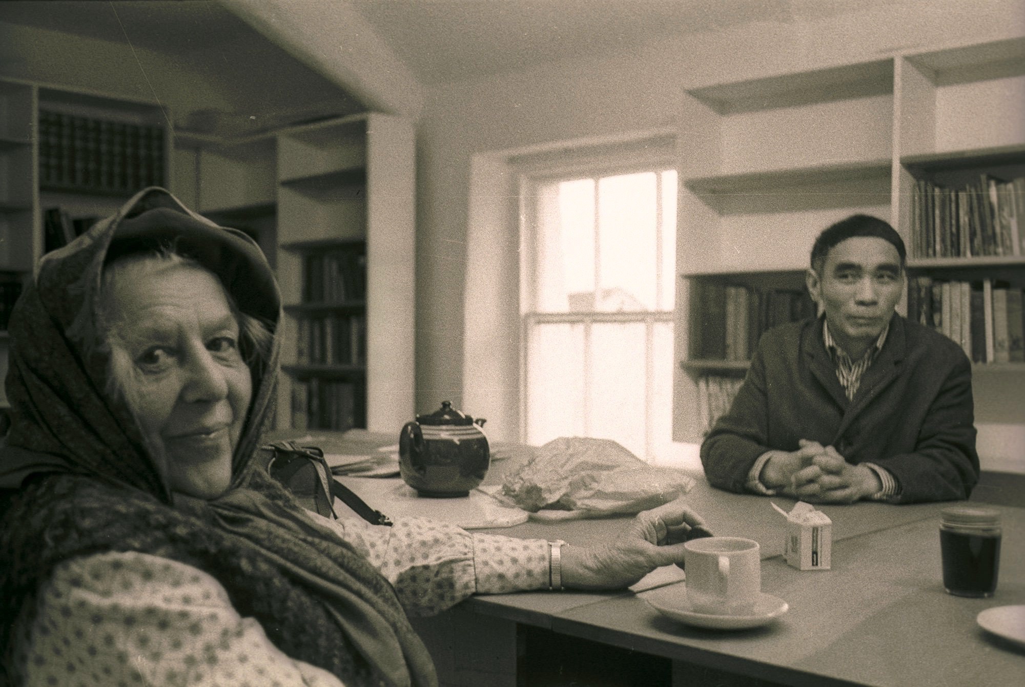 Black and white photograph. Two people sit at a table in a shelf-lined room. Closer to us, a pale-skinned woman’s head is wrapped in a scarf. She looks at us and smiles, one hand resting on the table. Across the table, a medium-skinned man sits with his fingers interlaced and slightly smiles, lips closed, at the woman.