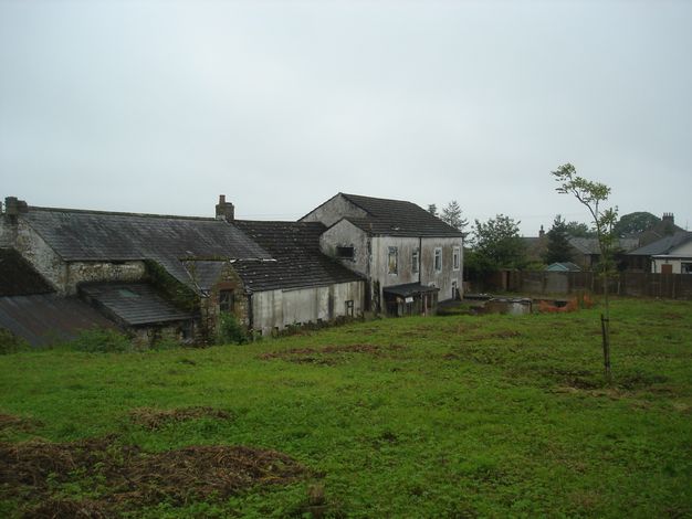Architecture. Grey-stained white structures are across a weedy lawn. A long, warehouse-like building is attached to what could be a two-storey house.